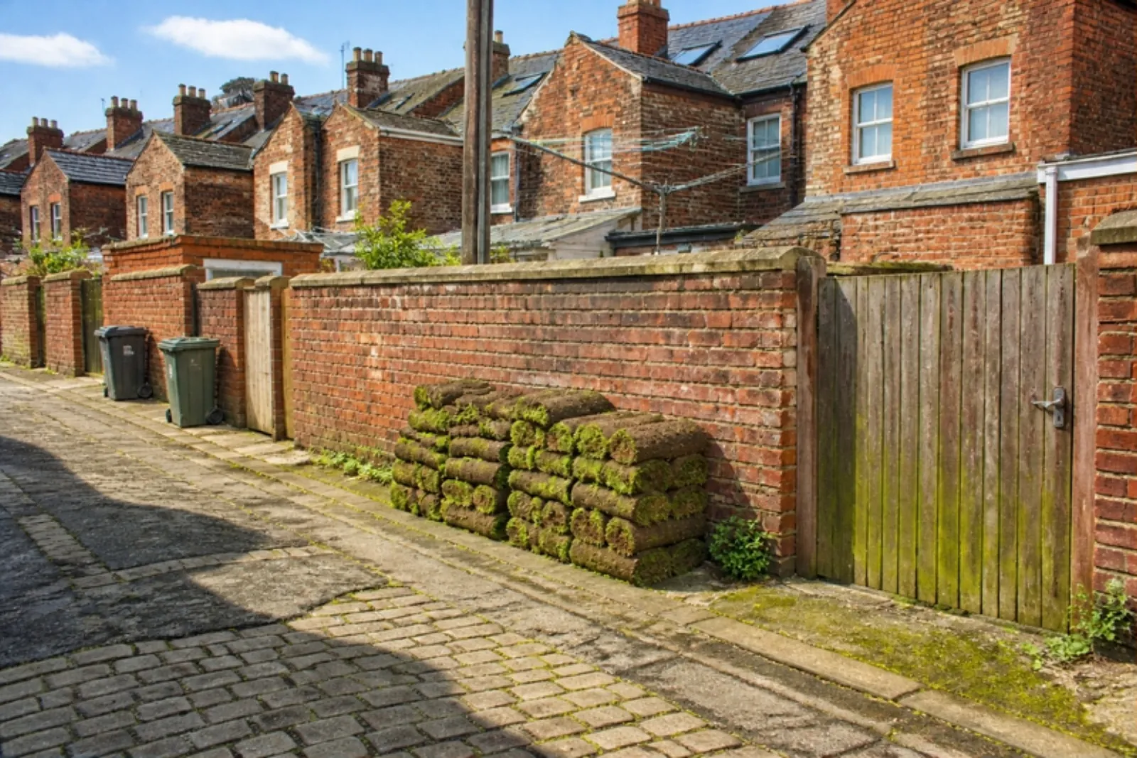 Rolls of turf stacked against a brick yard wall in a cobbled back lane behind red brick terraces